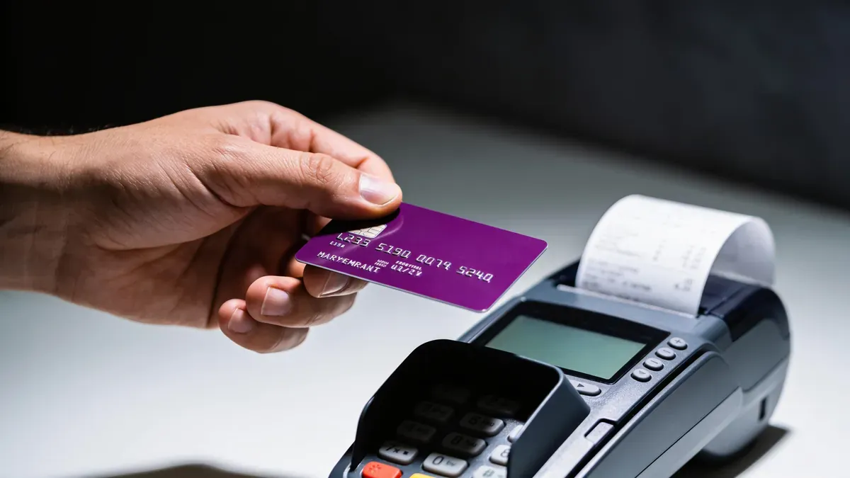 Mão segurando cartão de crédito pausada acima de terminal de pagamento, mostrando o momento da decisão antes de comprar