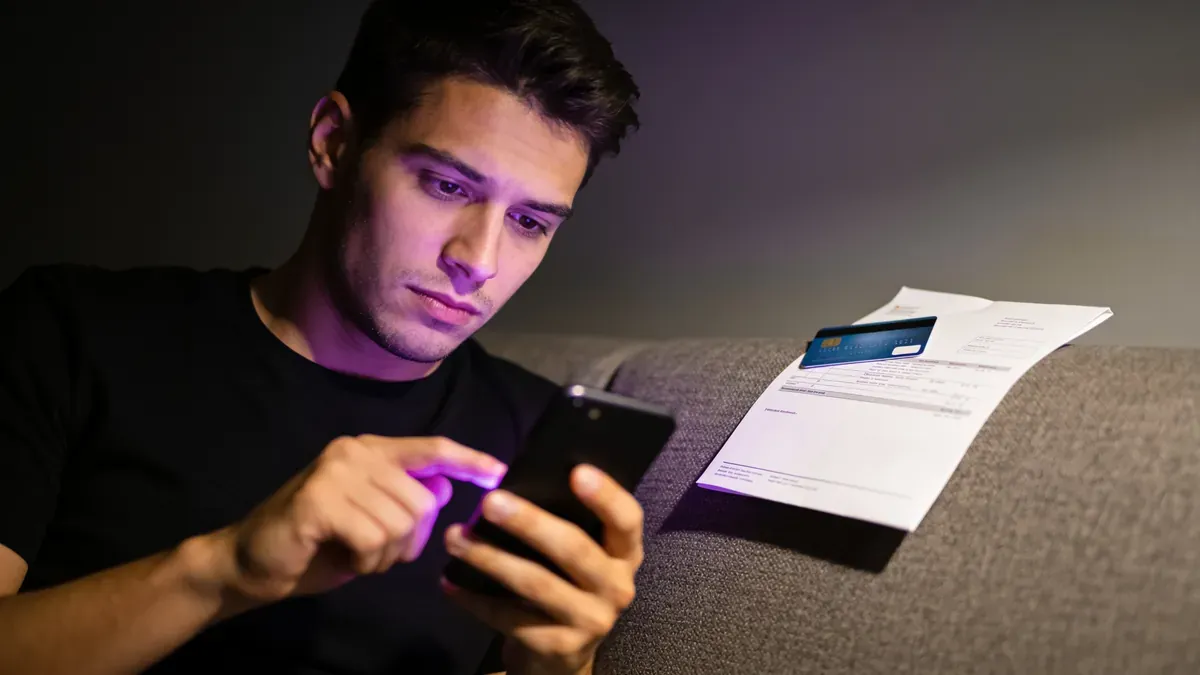Jovem adulto à noite reconhecendo uma chamada do banco no smartphone, com hesitação visível e uma fatura de cartão fechada ao lado