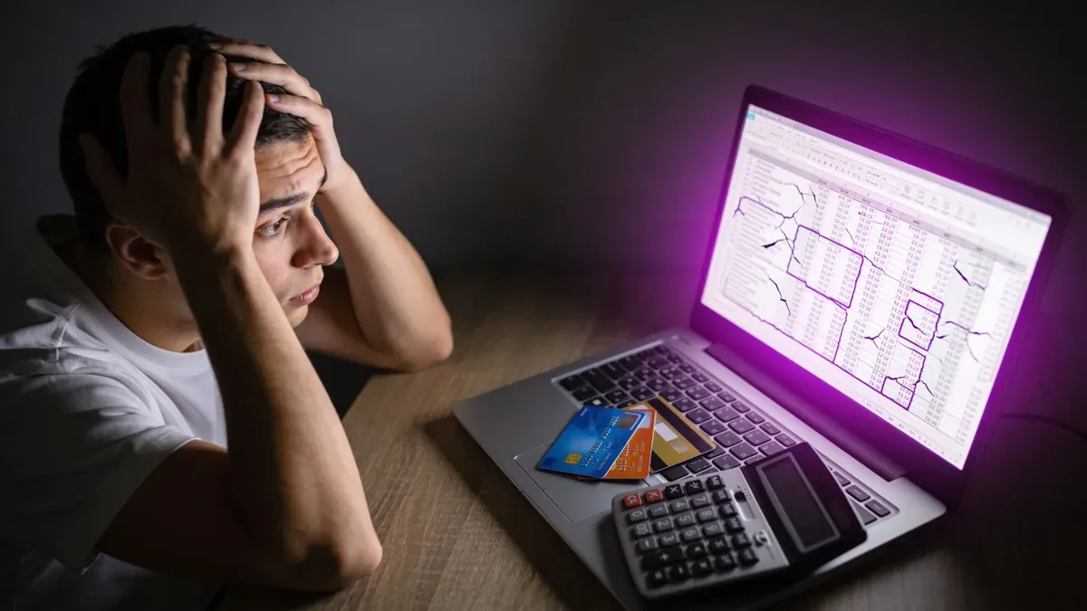 Rosto de jovem adulto com expressão de sobrecarga, olhando para tela de laptop com planilha densa à noite, cartões de crédito e calculadora sobre a mesa.