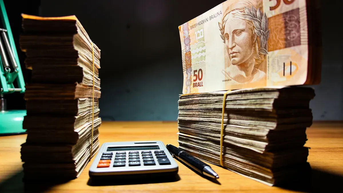 Duas pilhas de notas de real em tamanhos diferentes sobre uma mesa de madeira à noite, uma calculadora e caneta entre elas, iluminação dramática com acento verde esmeralda