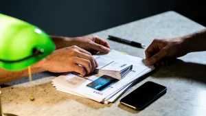 Mãos organizando contas e extrato de cartão numa mesa de cozinha, com caneta e smartphone ao lado, iluminação dramática com contraste e detalhe em verde.