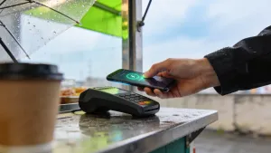 Mão tapping smartphone em pagamento mobile no counter de uma banca de rua, foco no gesto de aproximação, luz natural com accent verde-esmeralda do reflexo de awning urbano.