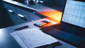 Computador portátil com planilha aberta e extrato do cartão impresso, noite, luz dramática em fundo neutro.