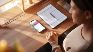 Mulher brasileira em seu home office decidindo entre planilha física e app, mão hesitante entre celular e caderno
