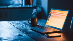 Laptop iluminado na mesa de madeira à noite com planilha de 5 colunas simples na tela, xícara de chá ao lado e celular deitado sobre a mesa.