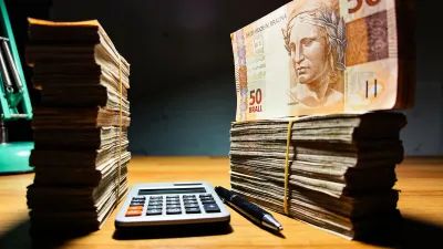 Duas pilhas de notas de real em tamanhos diferentes sobre uma mesa de madeira à noite, uma calculadora e caneta entre elas, iluminação dramática com acento verde esmeralda