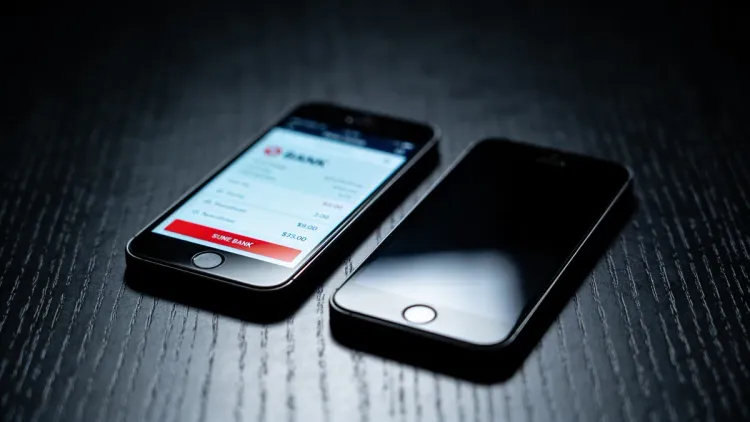 Dois celulares lado a lado sobre mesa escura, um com tela de app bancário iluminada, o outro apagado, mostrando o contraste de visibilidade entre as pessoas do casal.